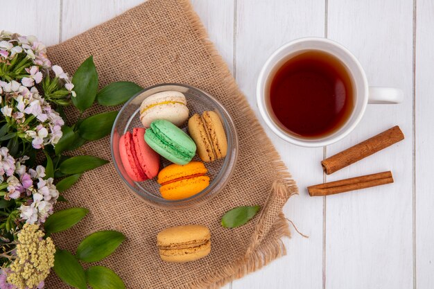 Vista superior de macarons coloridos em uma jarra com flores e uma xícara de chá com canela em uma superfície branca