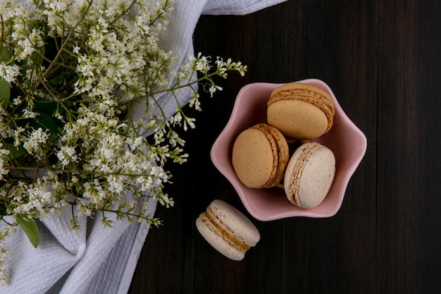 Vista superior de macarons em um pires com flores em uma toalha branca em uma superfície de madeira