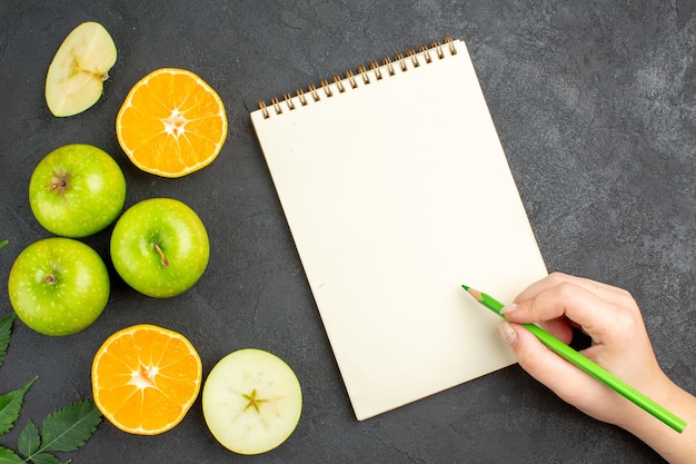 Vista superior de maçãs verdes frescas cortadas inteiras e picadas e laranjas cortadas com hortelã ao lado do caderno com caneta em fundo preto