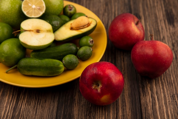 Vista superior de maçãs vermelhas frescas com frutas frescas, como maçãs verdes, limão, feijoas e abacates em um prato amarelo na parede de madeira