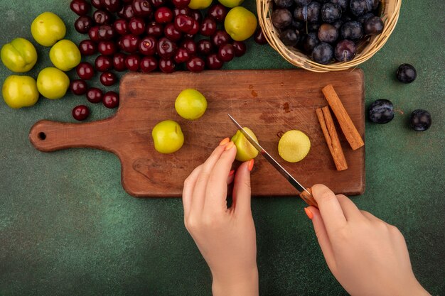 Vista superior de mãos femininas cortadas em pedaços de ameixa cereja verde com uma faca em uma placa de cozinha de madeira com cerejas vermelhas e abrunhos em um balde sobre um fundo verde