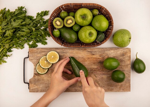 Vista superior de mãos femininas cortando abacate maduro com faca em uma placa de cozinha de madeira com limas com maçãs em um balde em um fundo branco