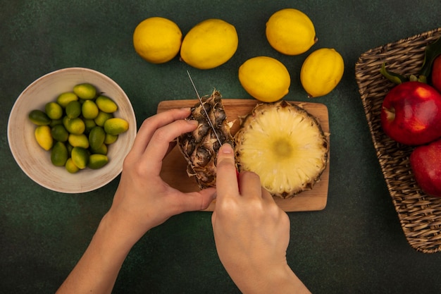 Vista superior de mãos femininas cortando abacaxi fresco em uma placa de cozinha de madeira com uma faca com kinkans em uma tigela com limões isolados em um fundo verde