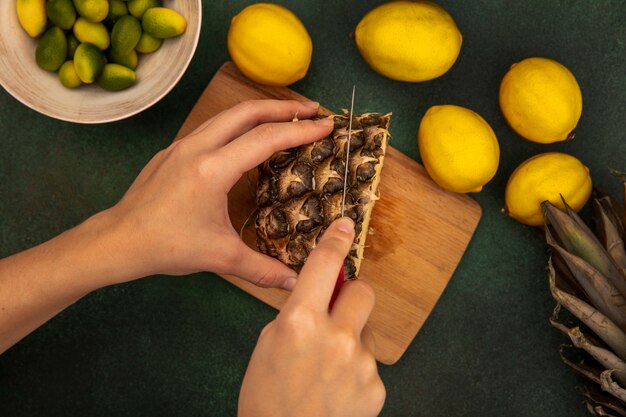 Vista superior de mãos femininas cortando abacaxi fresco em uma placa de cozinha de madeira com uma faca com kinkans em uma tigela com limões isolados em uma parede verde