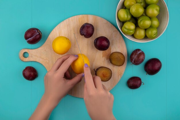 Vista superior de mãos femininas cortando nectacota com faca e pluots na placa de corte e pluots com tigela de ameixas no fundo azul