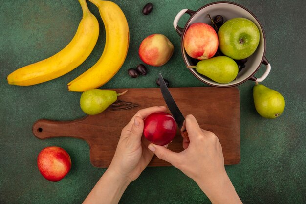 Vista superior de mãos femininas cortando pêssego com uma faca na tábua e banana pera uva maçã em fundo verde