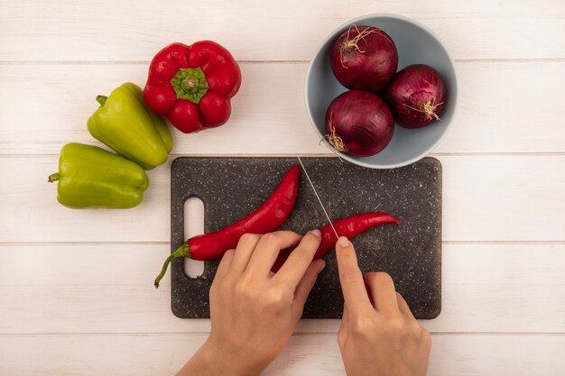 Vista superior de mãos femininas cortando pimenta em uma placa preta de cozinha com uma faca em uma parede de madeira branca