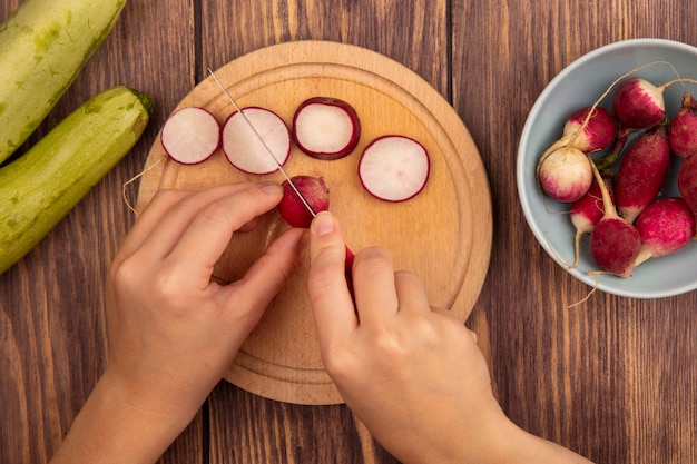 Vista superior de mãos femininas cortando rabanete em uma placa de cozinha de madeira com uma faca com rabanetes em uma tigela com abobrinhas isoladas em uma parede de madeira