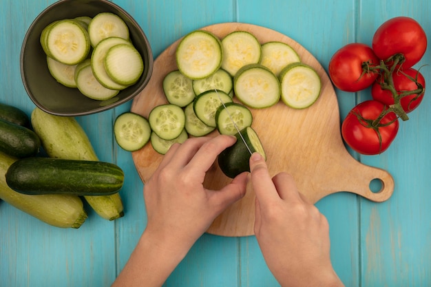 Vista superior de mãos femininas cortando um pepino fresco em uma mesa de madeira da cozinha com uma faca com tomates, pepinos e abobrinhas isoladas em uma parede de madeira azul