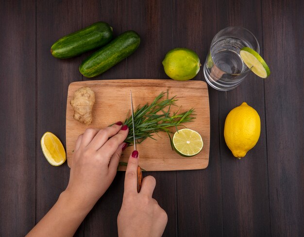 Vista superior de mãos femininas cortando verduras de estragão na mesa de madeira da cozinha com uma faca com limões na madeira