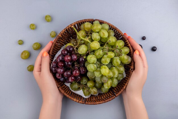 Vista superior de mãos femininas segurando uma cesta de uvas com bagas de uva em fundo cinza