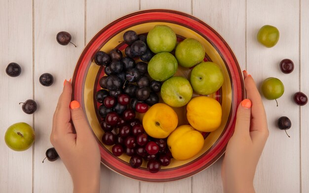 Vista superior de mãos femininas segurando uma tigela com frutas frescas, como pêssegos verdes cereja com ameixa cherriessweet em um fundo branco de madeira