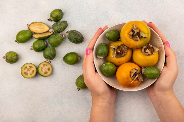 Foto grátis vista superior de mãos femininas segurando uma tigela de feijoas verdes maduras com caquis em uma superfície cinza