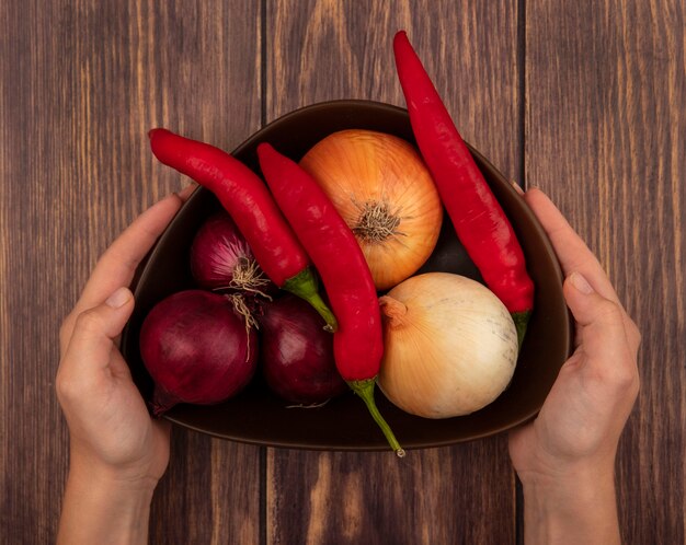 Vista superior de mãos femininas segurando uma tigela de legumes frescos, como cebola e pimentão, em uma superfície de madeira