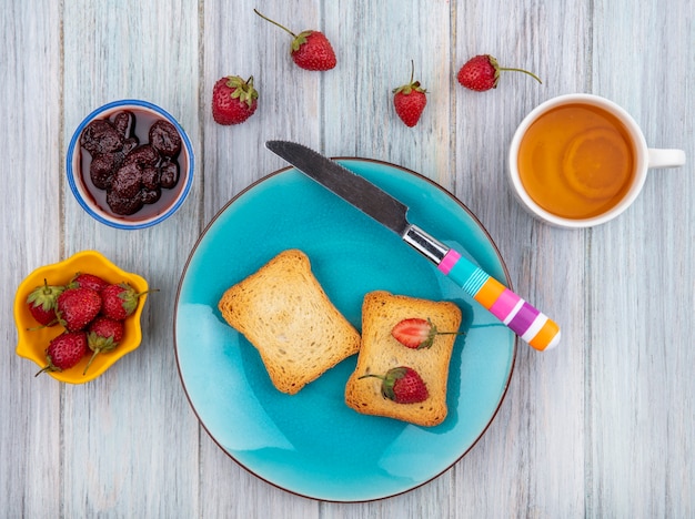 Vista superior de morango fresco em um pão torrado em um prato azul com faca com geléia de morango com morangos frescos com uma xícara de chá em um fundo cinza de madeira