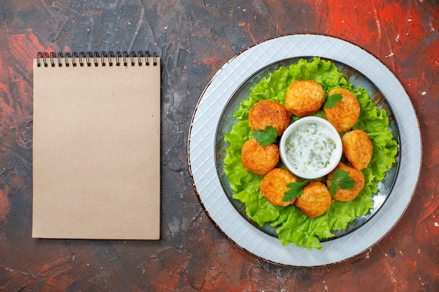 Vista superior de nuggets de frango, alface e molho, em um caderno de pratos na mesa escura