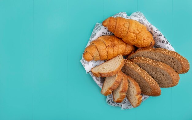 Vista superior de pães como baguete de sabugo fatiado e croissant em uma tigela sobre fundo azul com espaço de cópia