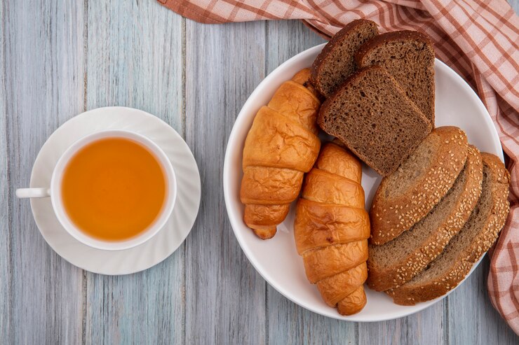 Foto grátis vista superior de pães como croissant fatiado de centeio e espiga marrom semeada em prato sobre pano xadrez e xícara de toddy quente em fundo de madeira