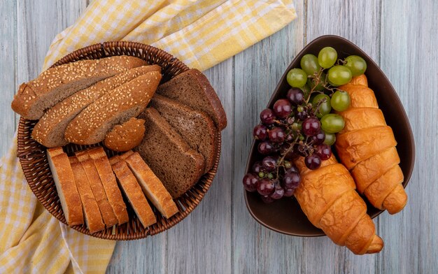 Vista superior de pães como fatias de centeio espiga marrom e crocante em uma cesta em tecido xadrez e uma tigela de croissants e uvas em fundo de madeira