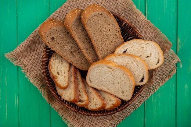 Vista superior de pães como fatias de sabugo marrom semeado e fatias de baguete em uma cesta em pano de saco sobre fundo verde