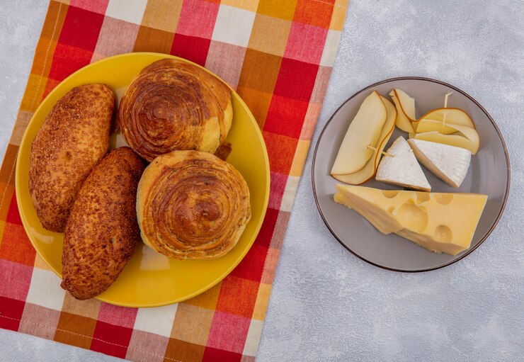 Foto grátis vista superior de pães frescos em um prato amarelo em um pano xadrez com diferentes tipos de queijo em um prato cinza em um fundo branco