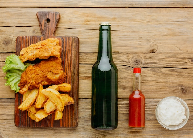 Vista superior de peixe e batatas fritas na tábua de cortar com garrafa de cerveja e ketchup Foto Premium