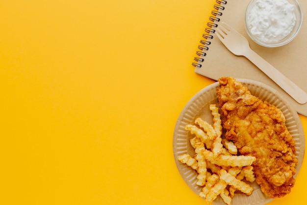 Vista superior de peixe e batatas fritas no prato com caderno e garfo