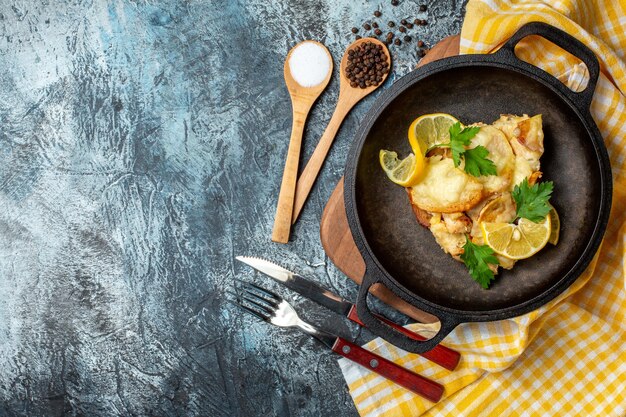 Vista superior de peixe frito em uma panela com especiarias diferentes de limão e salsa em colheres de madeira, garfo e faca em fundo cinza