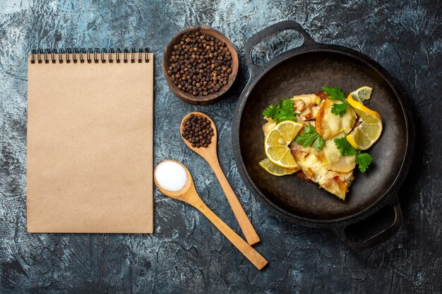 Vista superior de peixe frito na frigideira com especiarias de limão e salsa em colheres de madeira pimenta preta no caderno tigela no fundo cinza