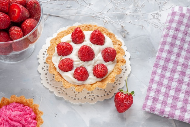 Vista superior de perto de um pequeno bolo com creme e morango vermelho fresco, bolo de creme rosa na mesa branca, bolo de frutas e biscoitos