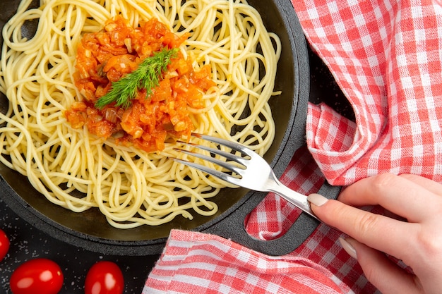 Vista superior de perto espaguete com molho no garfo de frigideira em tomate cereja feminino mão na mesa preta