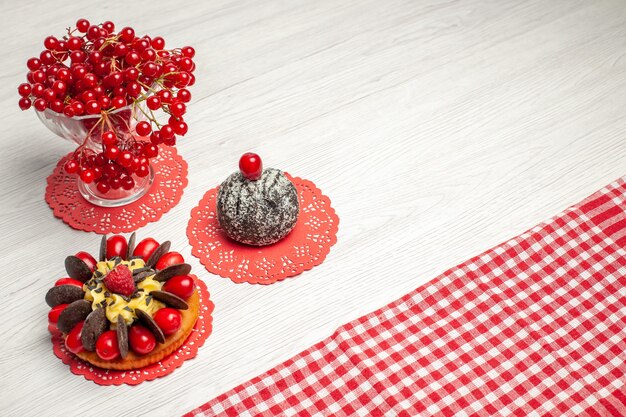 Vista superior de perto groselha em um bolo de frutas de cristal e bolo de cacau no guardanapo de renda oval vermelha e toalha de mesa quadriculada vermelho-branco na mesa de madeira branca