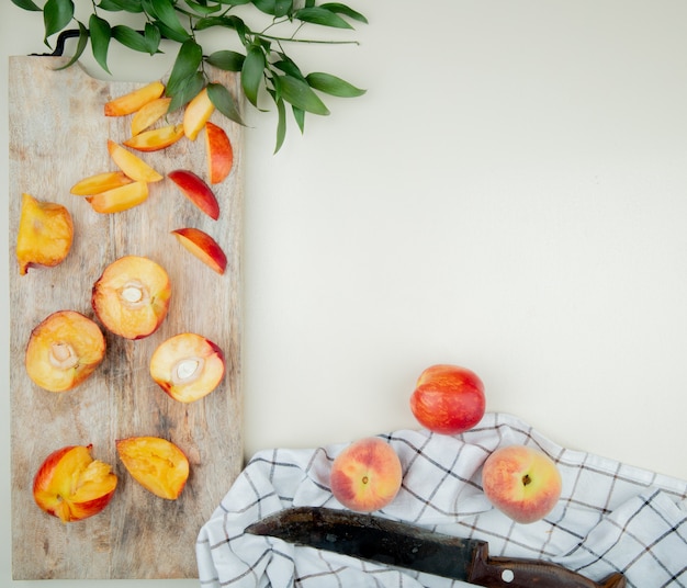 Vista superior de pêssegos cortados e fatiados na tábua com pêssegos inteiros e faca no pano na superfície branca, decorada com folhas com espaço de cópia