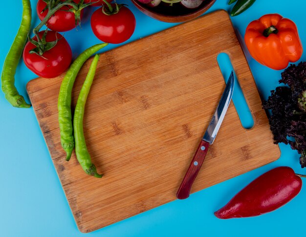 Vista superior de pimenta fresca em uma tábua de madeira com faca de cozinha e tomates maduros em azul