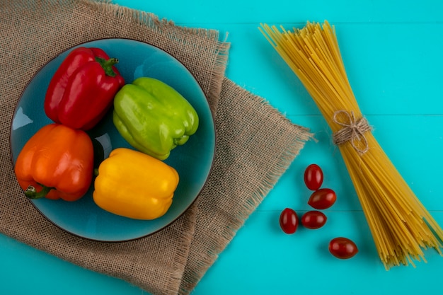 Vista superior de pimentões coloridos em um prato azul com massa crua e tomate cereja em uma superfície turquesa