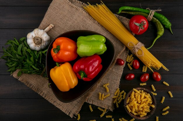 Vista superior de pimentões coloridos em uma tigela com espaguete cru e macarrão com alho e tomate em uma superfície de madeira