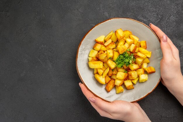 Vista superior de saborosas batatas fritas dentro do prato com verduras na superfície escura