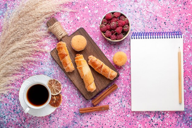 Vista superior de saborosas framboesas frescas dentro de um prato branco com pãezinhos de chá e canela na superfície rosa