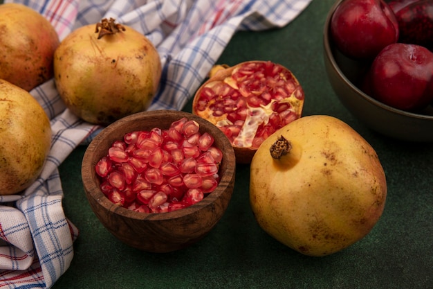 Vista superior de sementes de romã doce em uma tigela de madeira com pluots em uma tigela com faca com romãs isoladas em um pano xadrez em uma parede verde