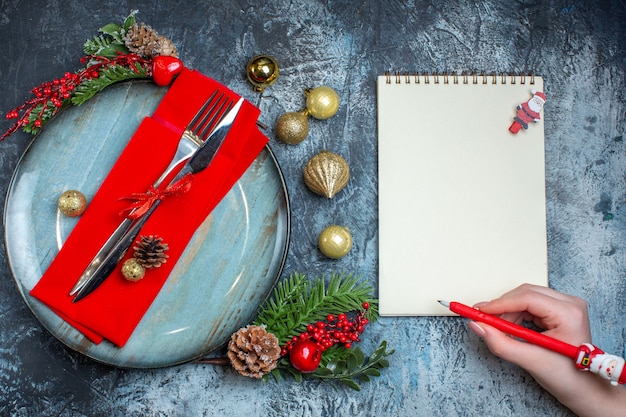Vista superior de talheres com fita vermelha em um guardanapo decorativo em um prato azul e acessórios de natal mão segurando a caneta no caderno em fundo escuro