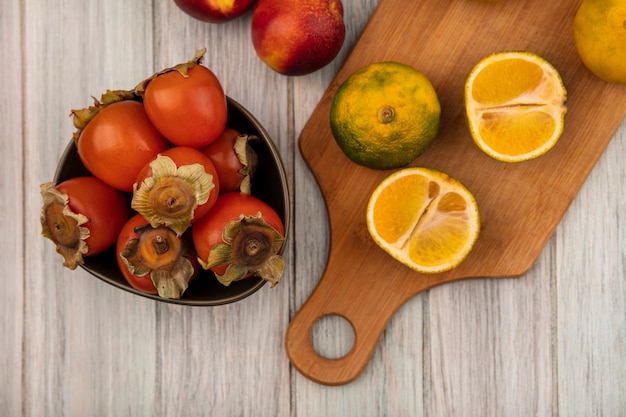 Vista superior de tangerinas saudáveis em uma placa de cozinha de madeira com caquis em uma tigela com pêssegos isolados em uma parede de madeira cinza