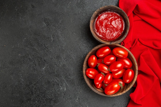 Vista superior de tigelas com tomate cereja e ketchup e toalha vermelha em fundo escuro