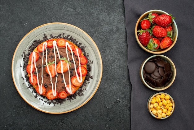 Vista superior de tigelas de bolo de avelã, morango e chocolate na toalha de mesa e bolo com chocolate e morango na mesa preta