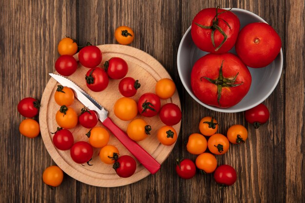 Vista superior de tomates cereja frescos vermelhos e laranja isolados em uma placa de cozinha de madeira com uma faca com tomates de tamanho grande em uma tigela na parede de madeira