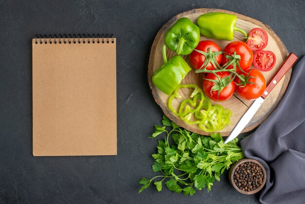 Vista superior de tomates frescos e pimentões verdes em um caderno de placa de madeira na superfície preta