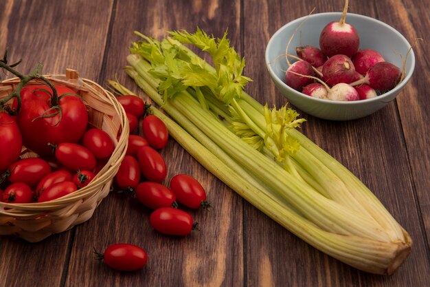 Vista superior de tomates saudáveis em um balde com rabanetes em uma tigela com aipo isolado em uma parede de madeira