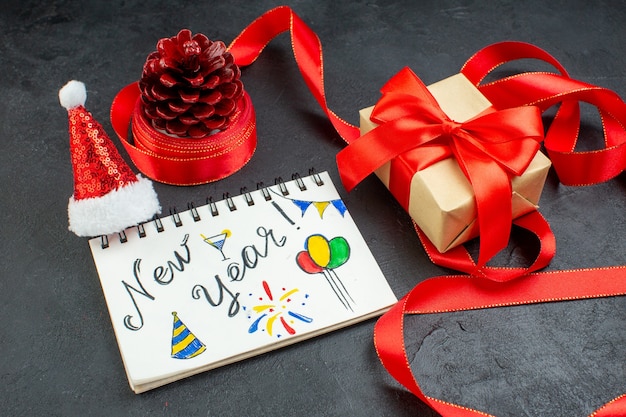 Vista superior de um cone de conífera de presente com fita vermelha e caderno com a escrita de ano novo e chapéu de papai noel lindo presente em fundo escuro