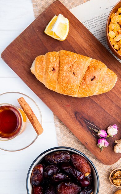 Vista superior de um croissant em uma tábua de madeira com copo de armudu de chá e tâmaras secas doces em uma tigela em madeira branca