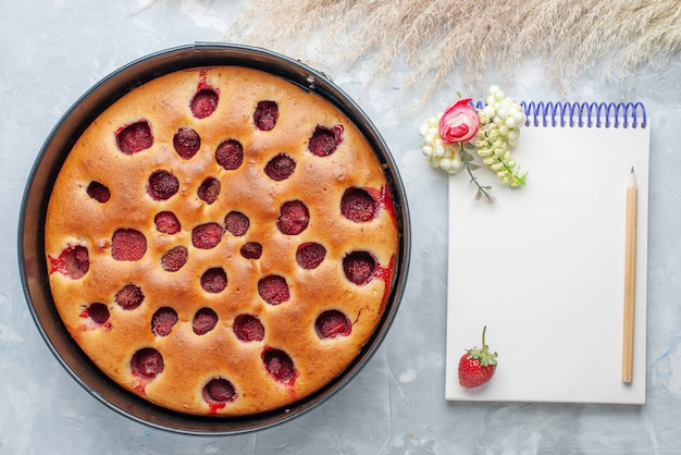 Foto grátis vista superior de um delicioso bolo de morango assado com morangos vermelhos frescos dentro com uma panela e um bloco de notas na mesa branca, bolo de biscoito doce de frutas