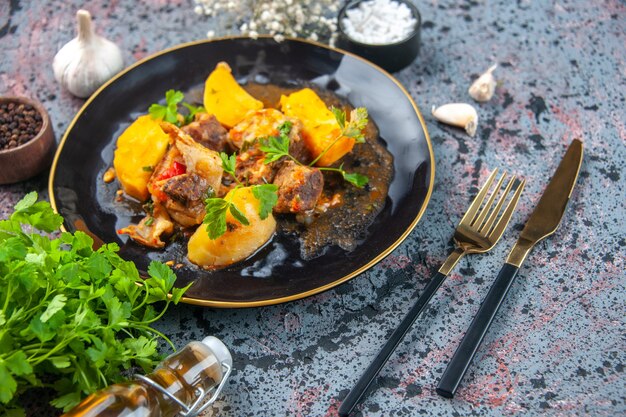 Vista superior de um jantar delicioso com batatas de carne servidas com verde em uma placa preta e especiarias de alho, talheres de flor de garrafa de óleo caídos em fundo de mistura de cores